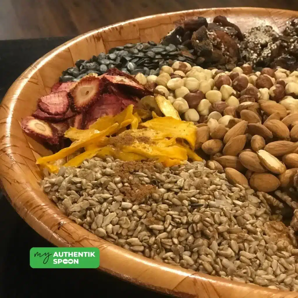 Ingredients for the trail mix recipe in wooden tray filled with activated sunflower seeds, dried mango, almonds, hazelnuts, strawberries, and other trail mix components.