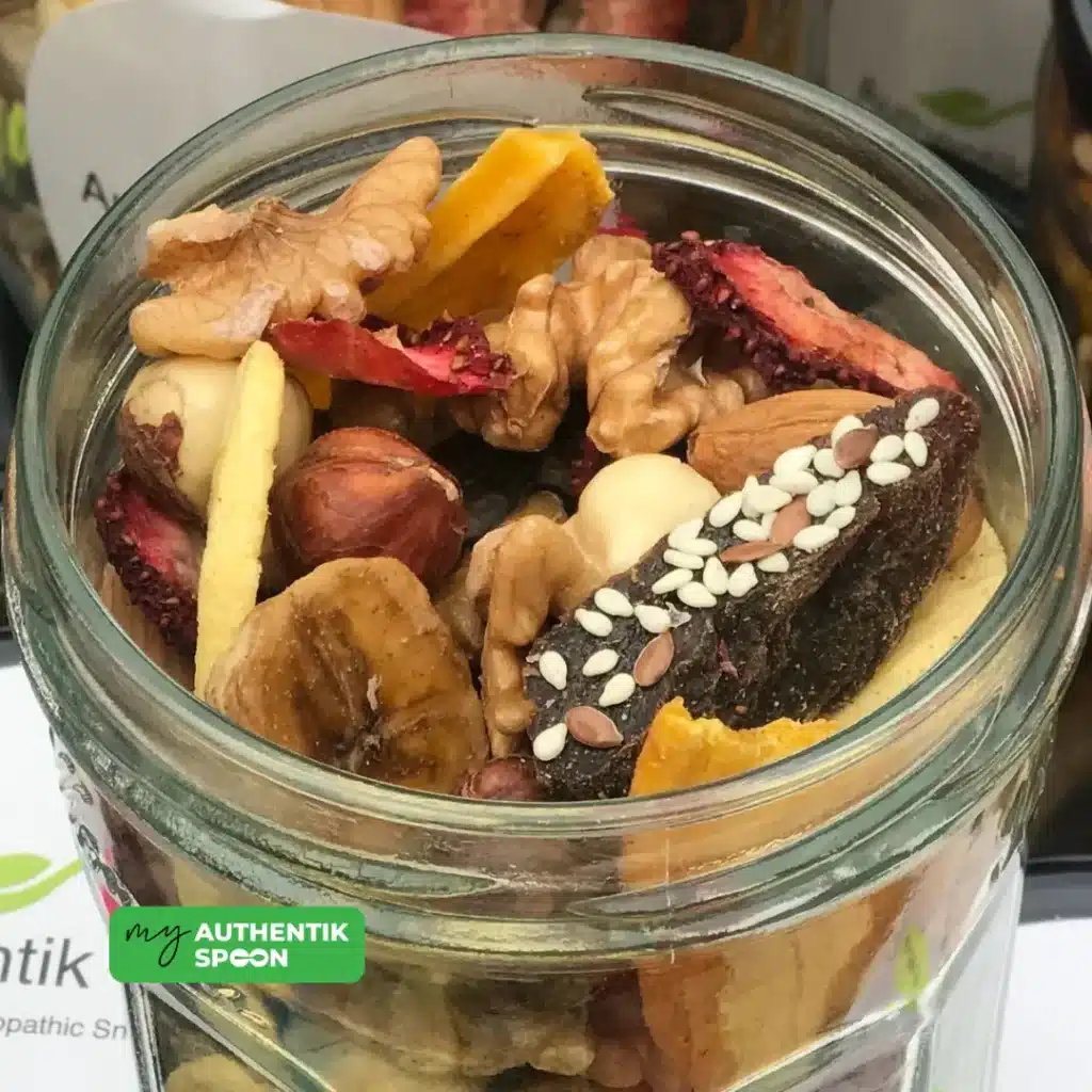 Glass jar filled with activated trail mix including banana chips, walnuts, hazelnuts, strawberries, and sesame-covered bites.