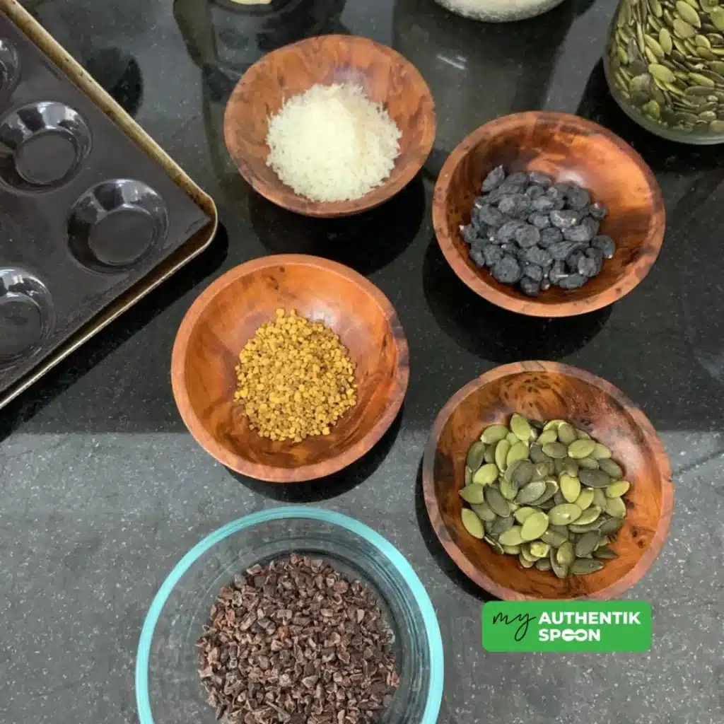 Ingredients for raw chocolate bites including cacao nibs, pumpkin seeds, raisins, bee pollen, shredded coconut, and nut butter